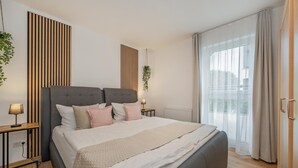 Ein modernes Schlafzimmer mit einem großen Bett, Nachttischen, einer hängenden Pflanze und Blick ins Freie durch ein Fenster.