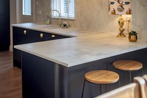 A kitchen with a marble countertop, two wooden stools, a gold decorative item, and a window with blinds. - Beachcomb Cottage | Elie (Elie)