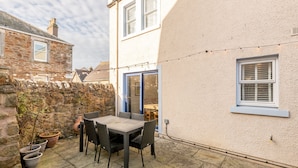 A patio with a table and chairs, a stone wall, and a building with a window. - Beachcomb Cottage | Elie (Elie)