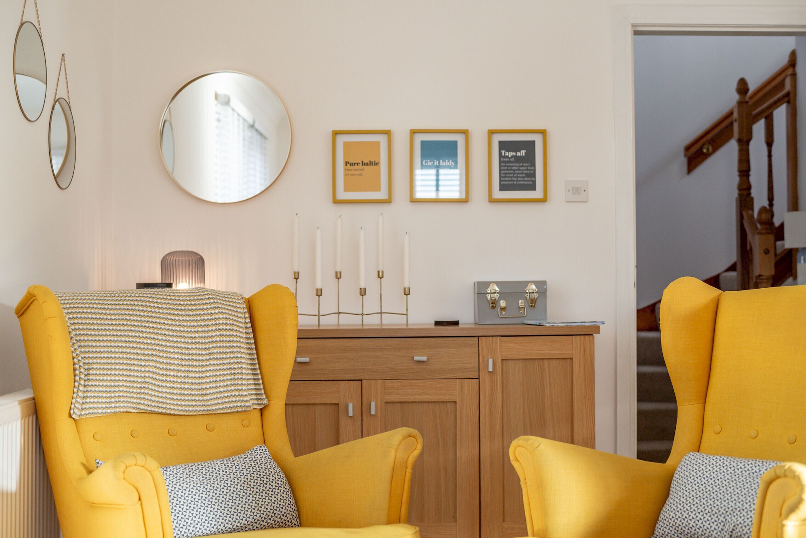Een kamer met twee gele fauteuils, een houten kast, een spiegel en drie ingelijste borden aan de muur.