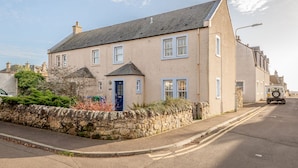 A two-story house with a stone wall, a blue door, and a parked vehicle on the street. - Beachcomb Cottage | Elie (Elie)