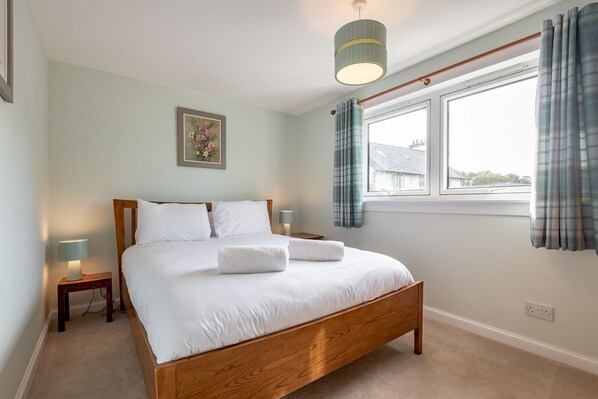 A neatly made bed with white linens and two towels, a bedside lamp, a framed picture on the wall, and a window with curtains.