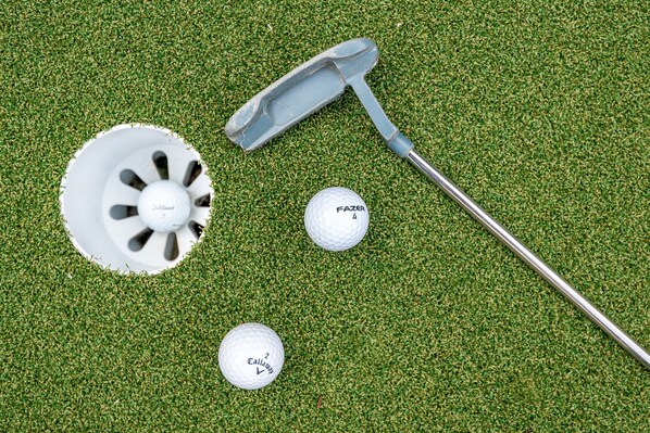 Yksi golfmaila, kaksi golfpalloa ja putteri vihreällä alustalla.