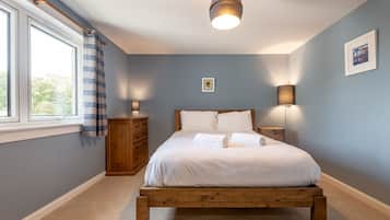 A bedroom with a wooden bed, a nightstand, a dresser, a window with curtains, and a wall-mounted lamp.