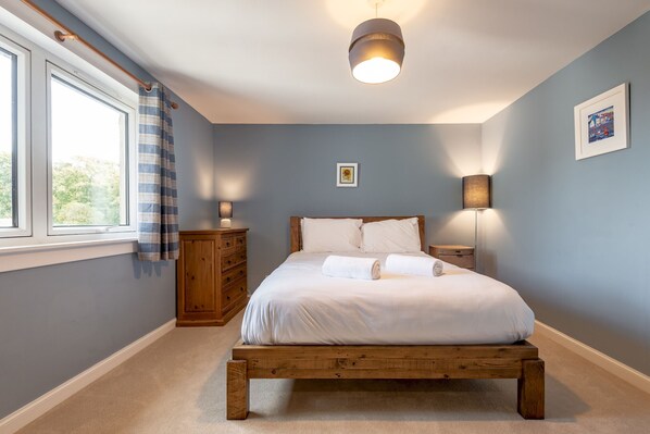 A bedroom with a wooden bed, a nightstand, a dresser, a window with curtains, and a wall-mounted lamp.