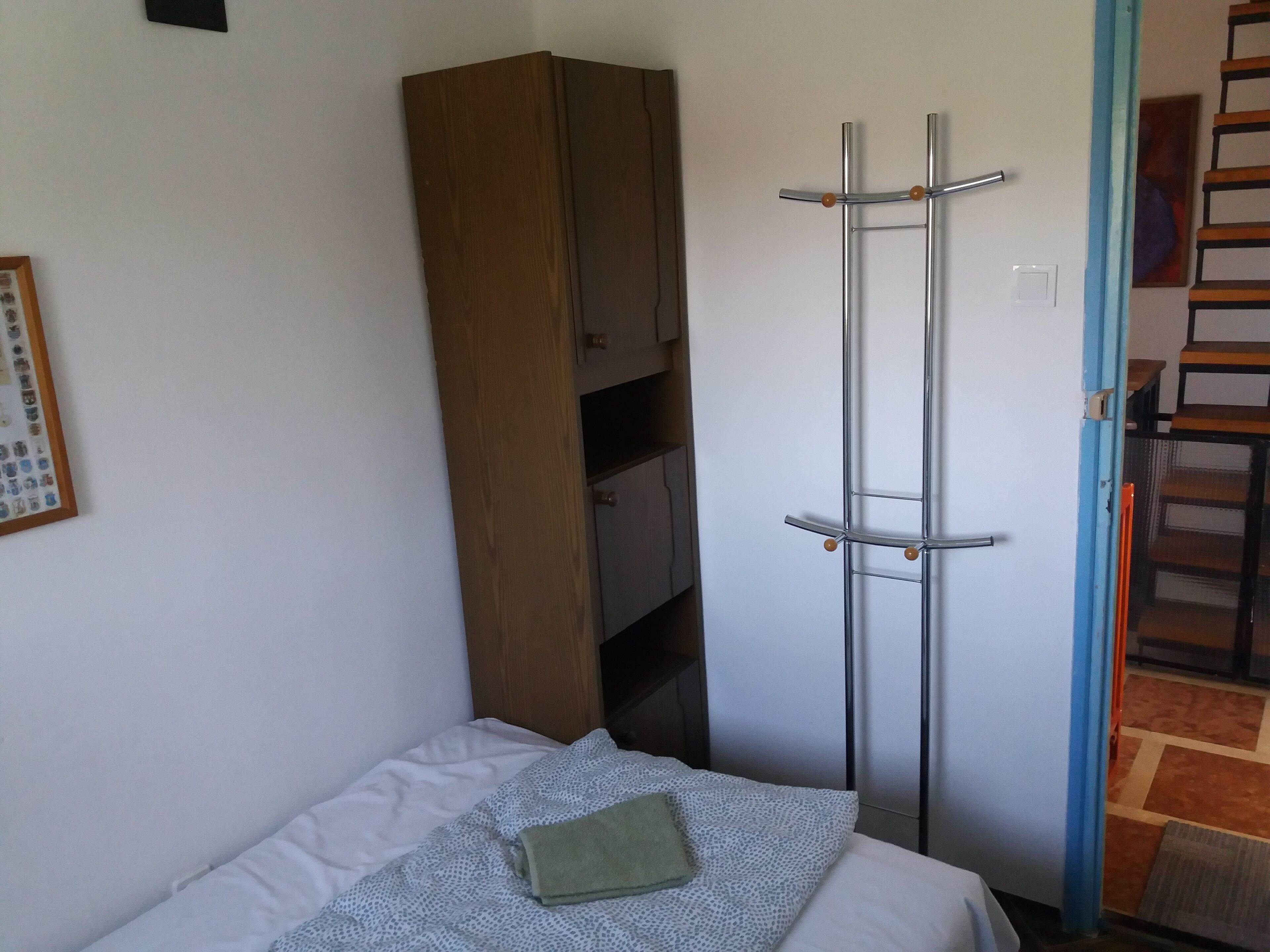 A small bedroom with a single bed, a wooden wardrobe, a metal coat rack, and a door leading to a staircase.