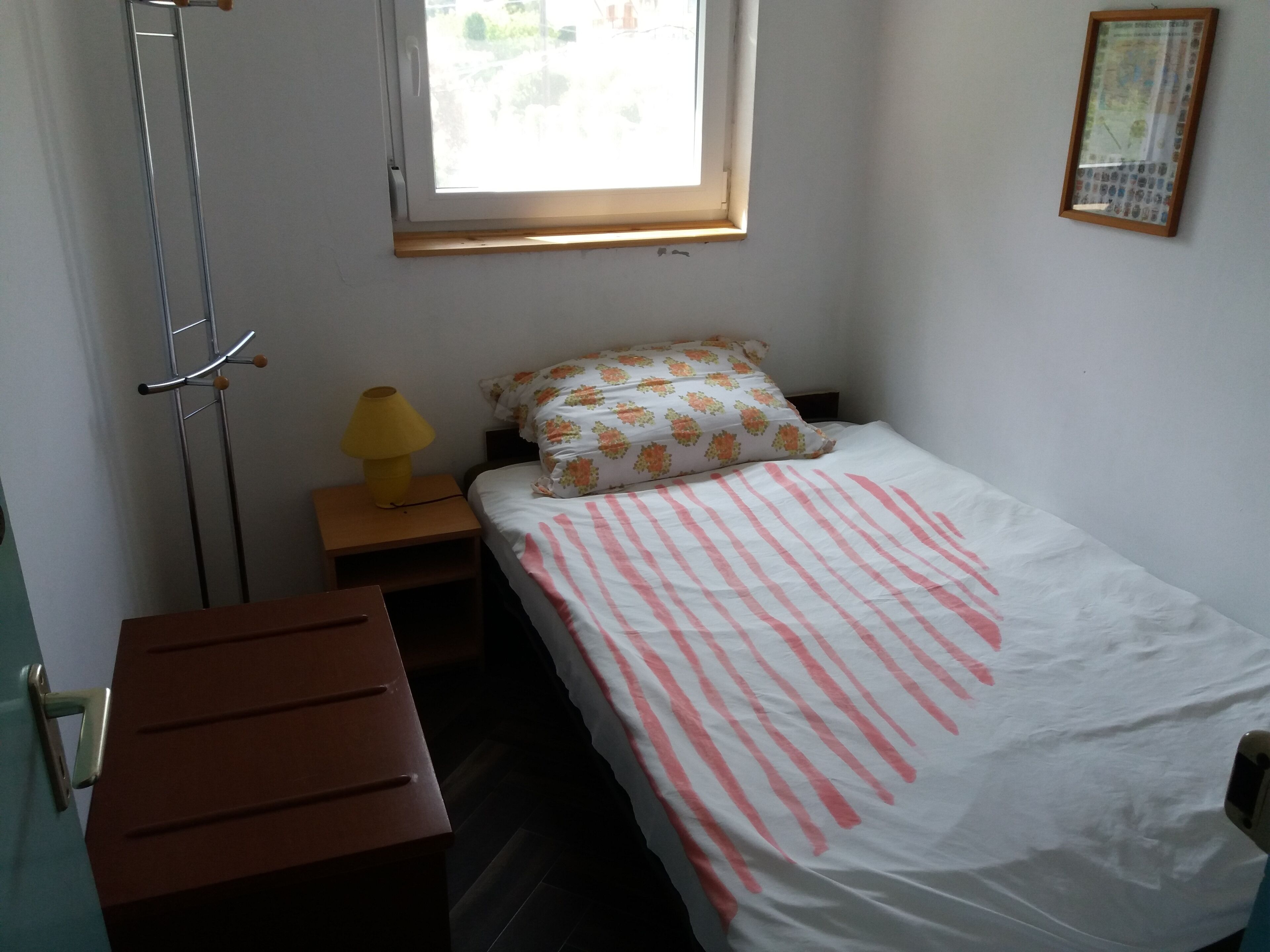 Ein kleines, ordentliches Zimmer mit einem Einzelbett, einem Nachttisch mit Lampe, einer Holzbank und einem Fenster mit Blick auf Grünflächen.