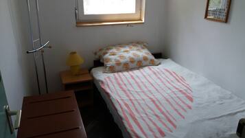 Ein kleines, ordentliches Zimmer mit einem Einzelbett, einem Nachttisch mit Lampe, einer Holzbank und einem Fenster mit Blick auf Grünflächen.