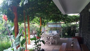 A covered outdoor dining area with a table and chairs, surrounded by greenery and plants.