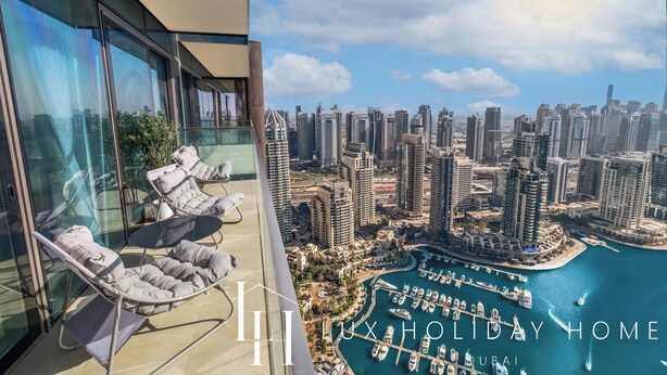 Un edificio de gran altura con un balcón que cuenta con sillas acolchadas y barandilla de vidrio. La vista muestra un puerto deportivo con barcos y un paisaje urbano con edificios altos.