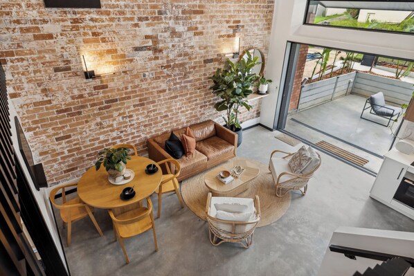 A loft-style living area with exposed brick walls, a round dining table, a leather sofa, and a view of the outdoor balcony.