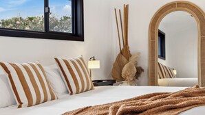 Un dormitorio moderno con una cama grande, almohadas a rayas, una lámpara de noche, una planta decorativa y un espejo.