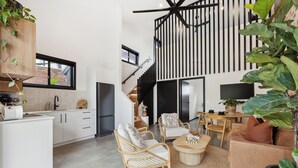 Una cocina moderna con armarios blancos, un refrigerador negro y una isla de madera. Hay una zona de comedor con sillas de mimbre y una mesa de centro. Una planta grande está en la esquina, y una escalera conduce a un nivel superior.