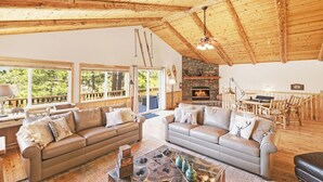 Uma sala de estar ampla, com dois sofás, lareira, vigas de madeira no teto e grandes janelas com vista para o verde.