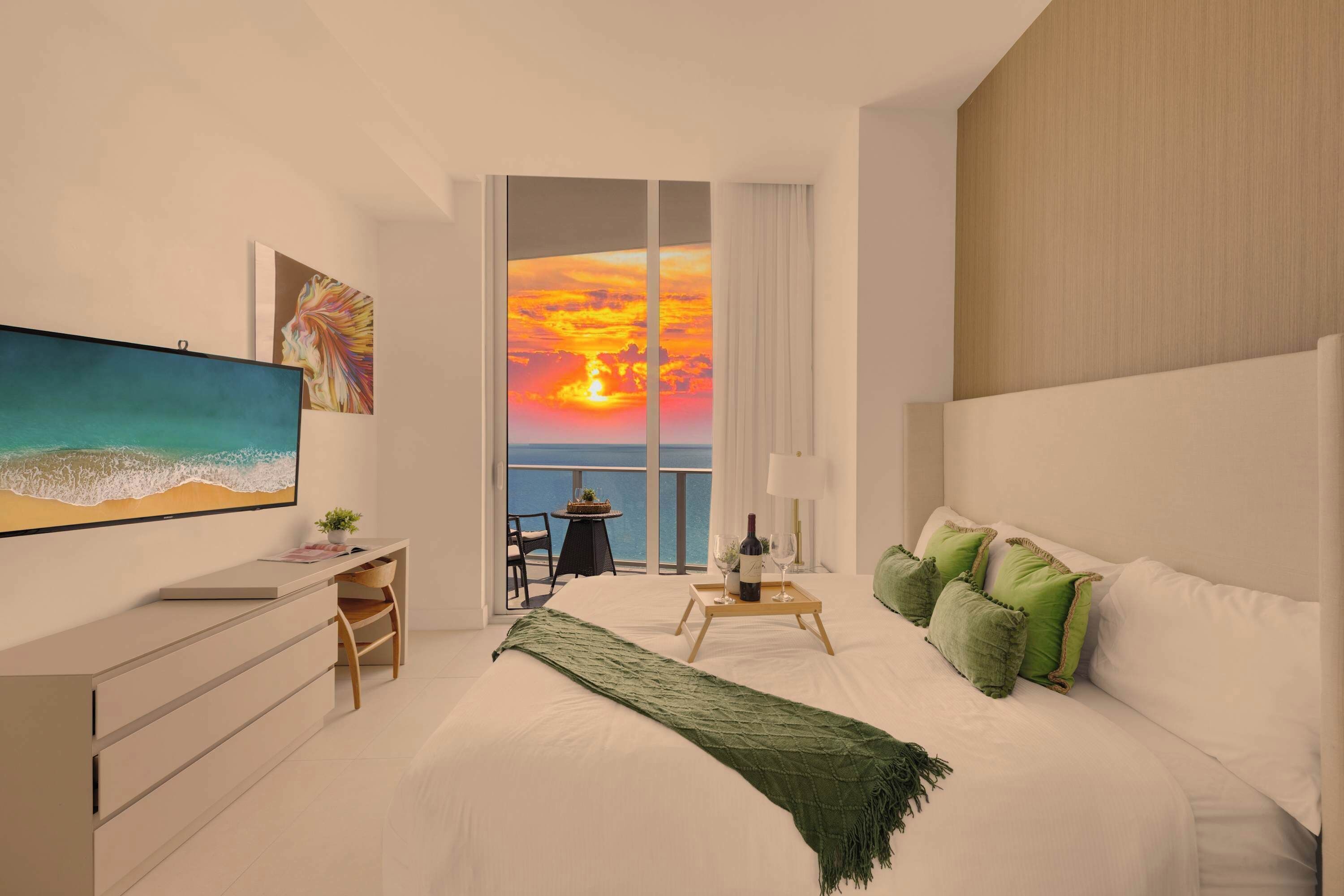 A modern hotel room with a large bed, a desk, a TV, and a balcony with a sunset view.