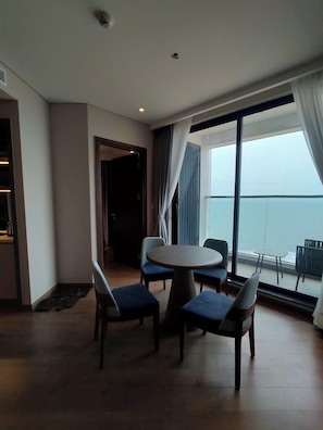 A modern hotel room with a round dining table and four chairs, a large window with a view of the sea, and a balcony with outdoor seating.