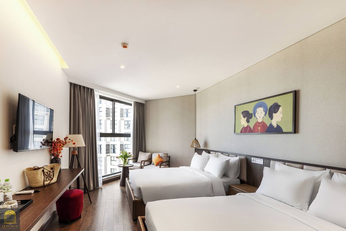 Ein Hotelzimmer mit zwei Betten, einem Schreibtisch mit Lampe, einem Fernseher und einem Fenster mit Vorhängen.