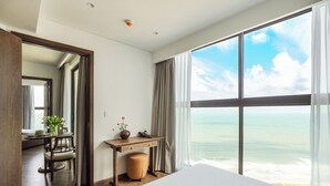 A hotel room with a large window offering a view of the ocean, a wooden desk with a vase of flowers, and a bed with white linens. - Apec Mandala Resort - Mui Ne (Mui Ne)