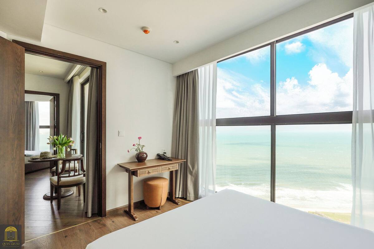 Ein Hotelzimmer mit großem Fenster und Meerblick, einem Holztisch mit Blumenvase sowie einem Bett mit weißer Bettwäsche.