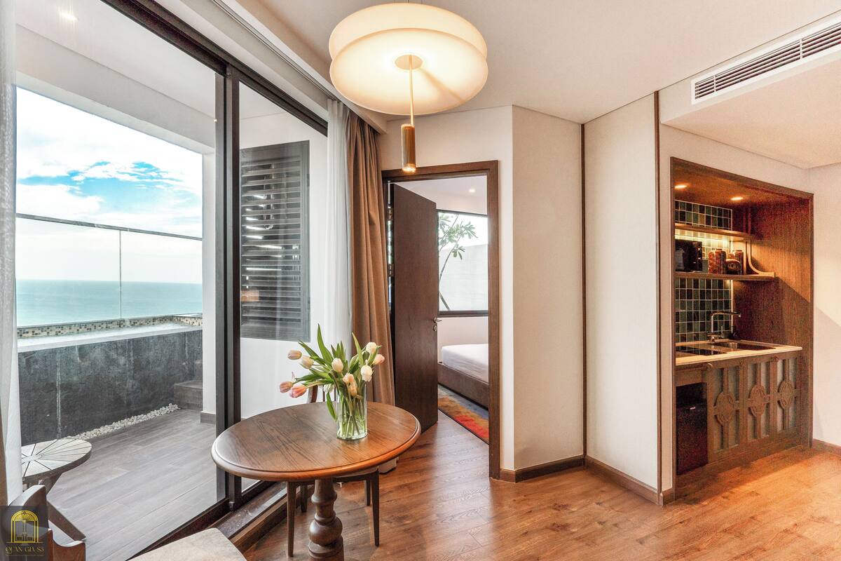Ein modernes Hotelzimmer mit Holzboden, einem runden Esstisch mit Blumenvase, einem großen Fenster mit Meerblick und einer Küchenzeile inklusive Spüle und Schränken.