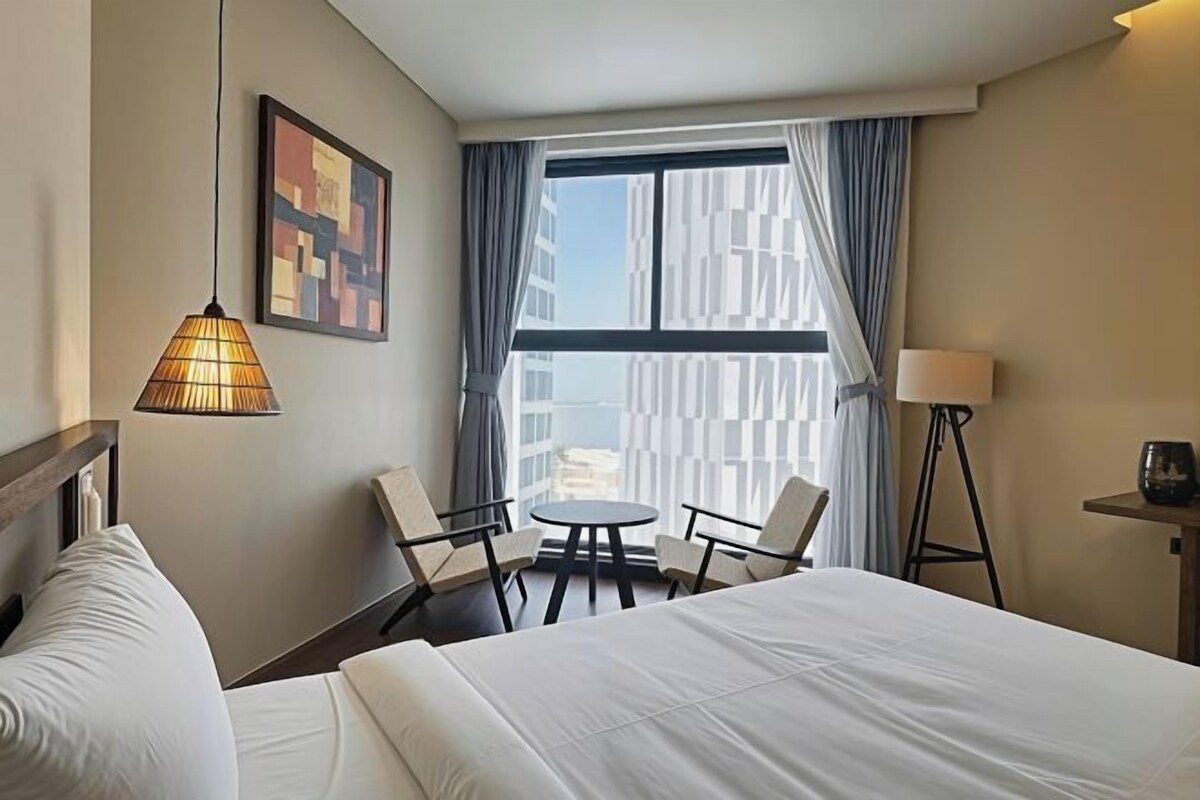 A hotel room with a bed, a table with chairs, a lamp, a painting, and a view through a window.