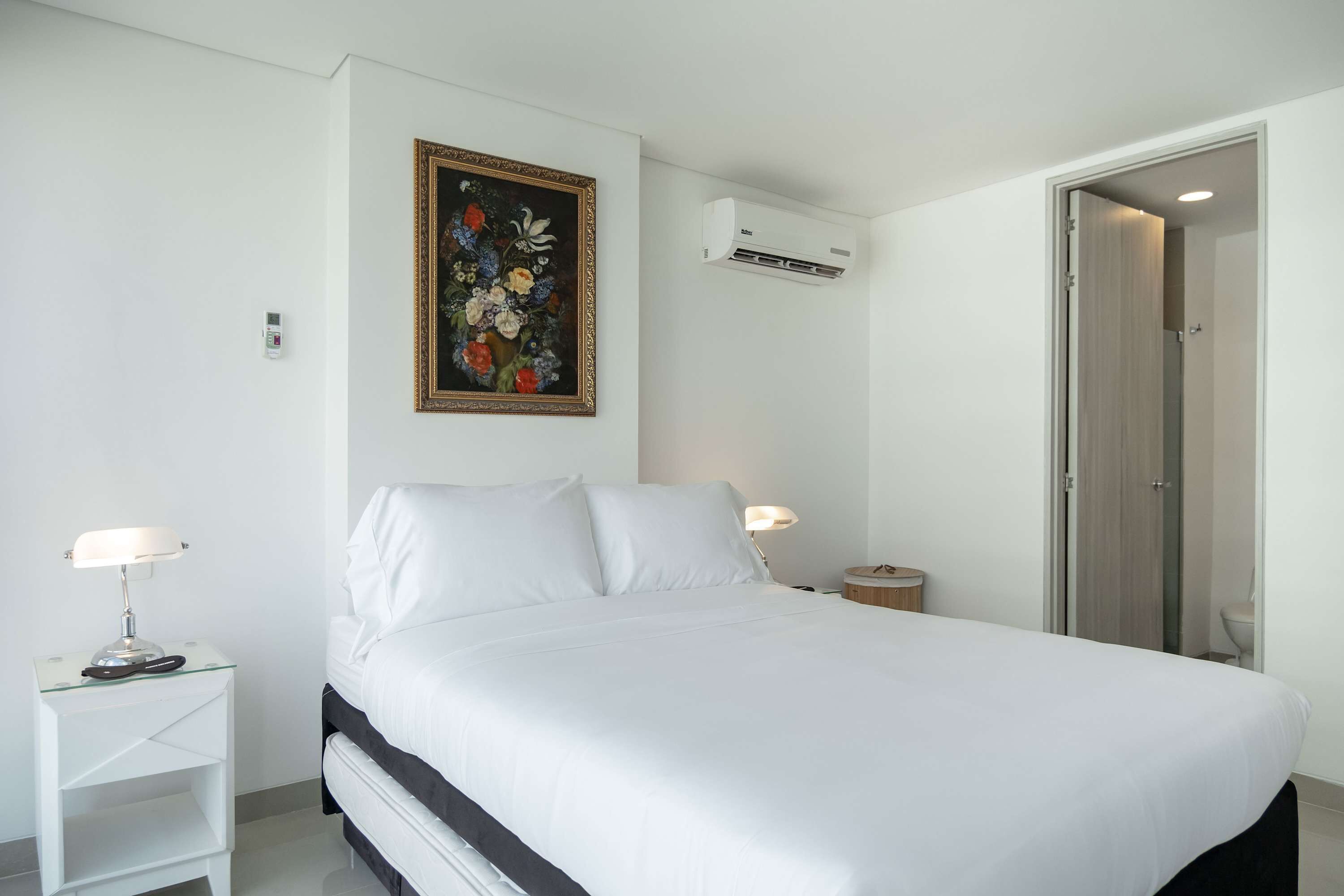 Un lit bien fait, avec du linge de lit blanc, une table de chevet avec une lampe, un climatiseur fixé au mur et une porte donnant sur une salle de bain.
