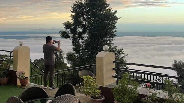 A terrace with wicker chairs, potted plants, and a person taking a photo. - The Unmad Satovari (Dharamshala)
