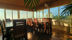 A dining area with a large table and chairs, large windows, and a view of the outdoors. - The Unmad Satovari (Dharamshala)