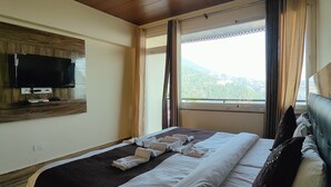 A hotel room with a bed, a flat-screen TV mounted on a wooden panel, a window with curtains, and a view of a mountainous landscape. - The Unmad Satovari (Dharamshala)