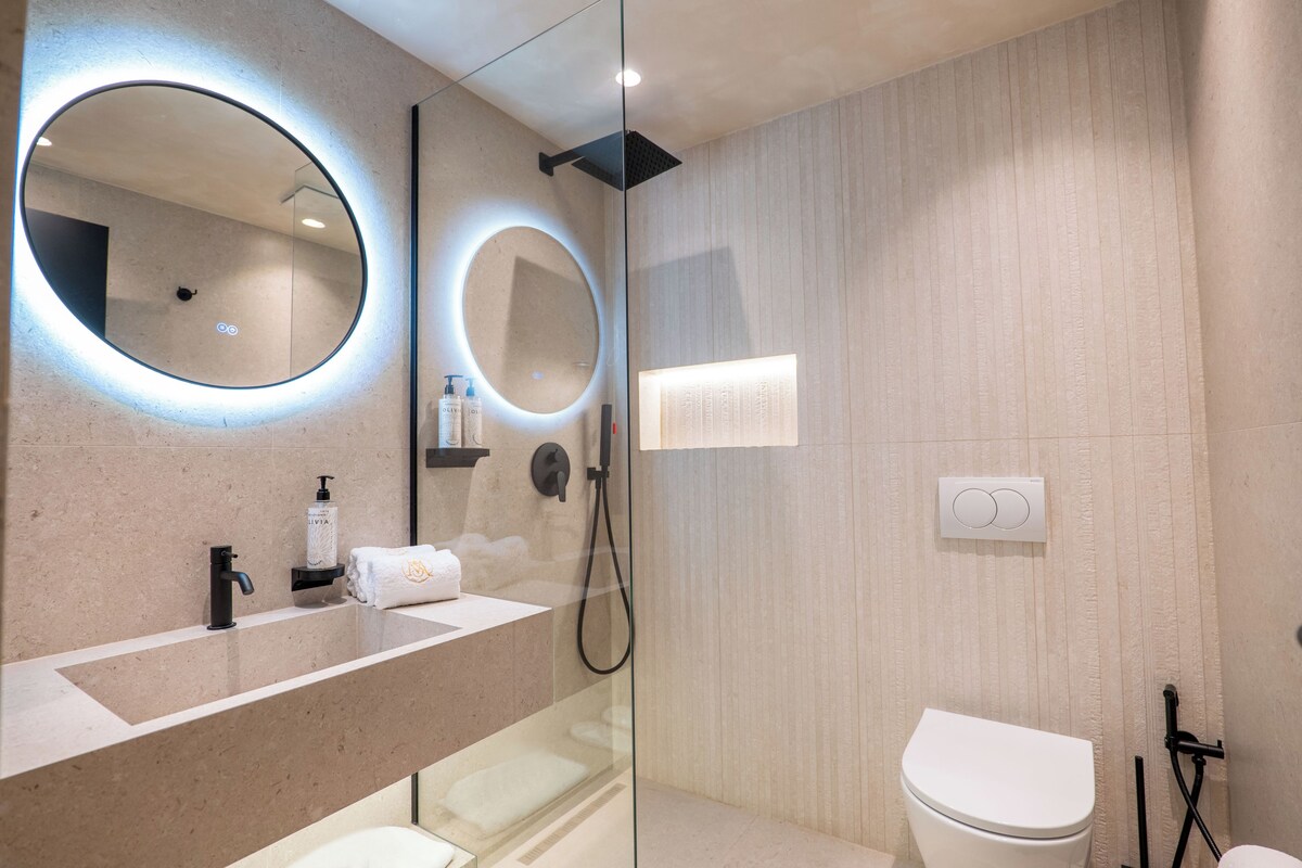 A modern bathroom with a glass-enclosed shower, a round mirror with underglow, a white toilet, and a beige countertop with a faucet and soap dispenser.