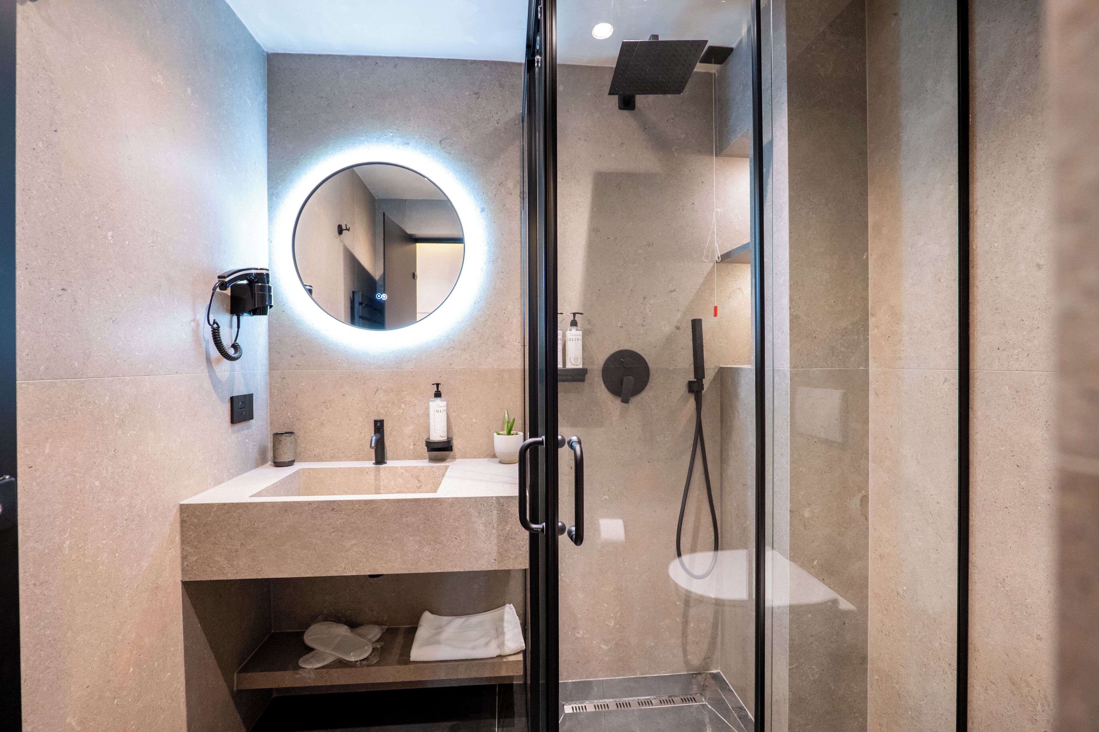 Une salle de bain moderne avec une douche à parois de verre, un grand miroir rond avec éclairage intégré, un sèche-cheveux et un lavabo avec un porte-serviettes.