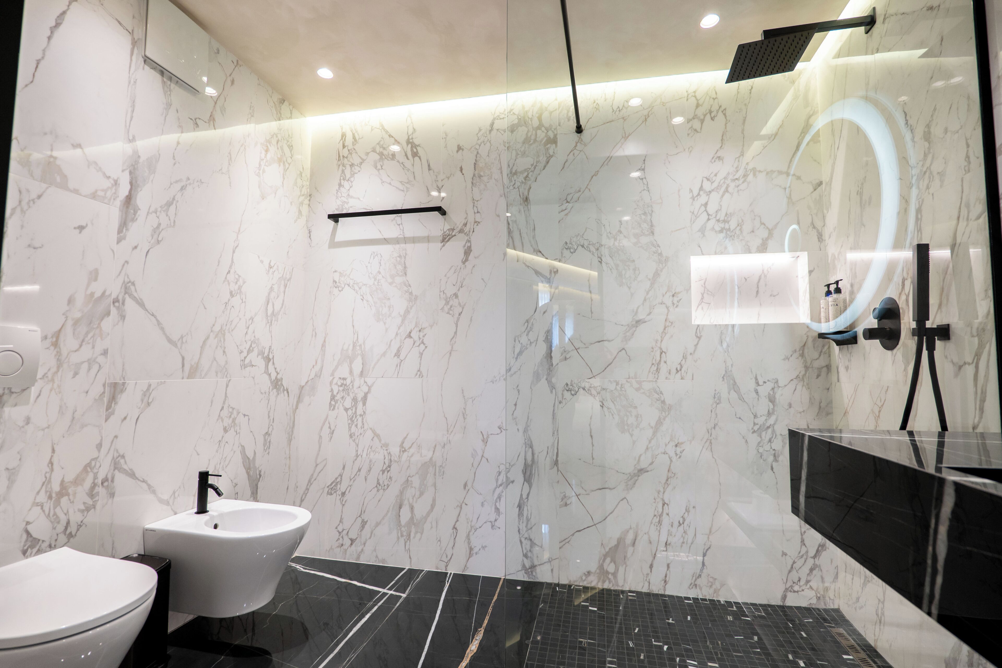 Un baño moderno con pared de mármol, un lavamanos negro, un bidé y una cabina de ducha de vidrio.