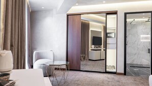 A hotel room with a bed, a desk, a chair, a small table, a TV, a bathroom with a shower, and a mirror. - Mattis Hotel (Tirana)