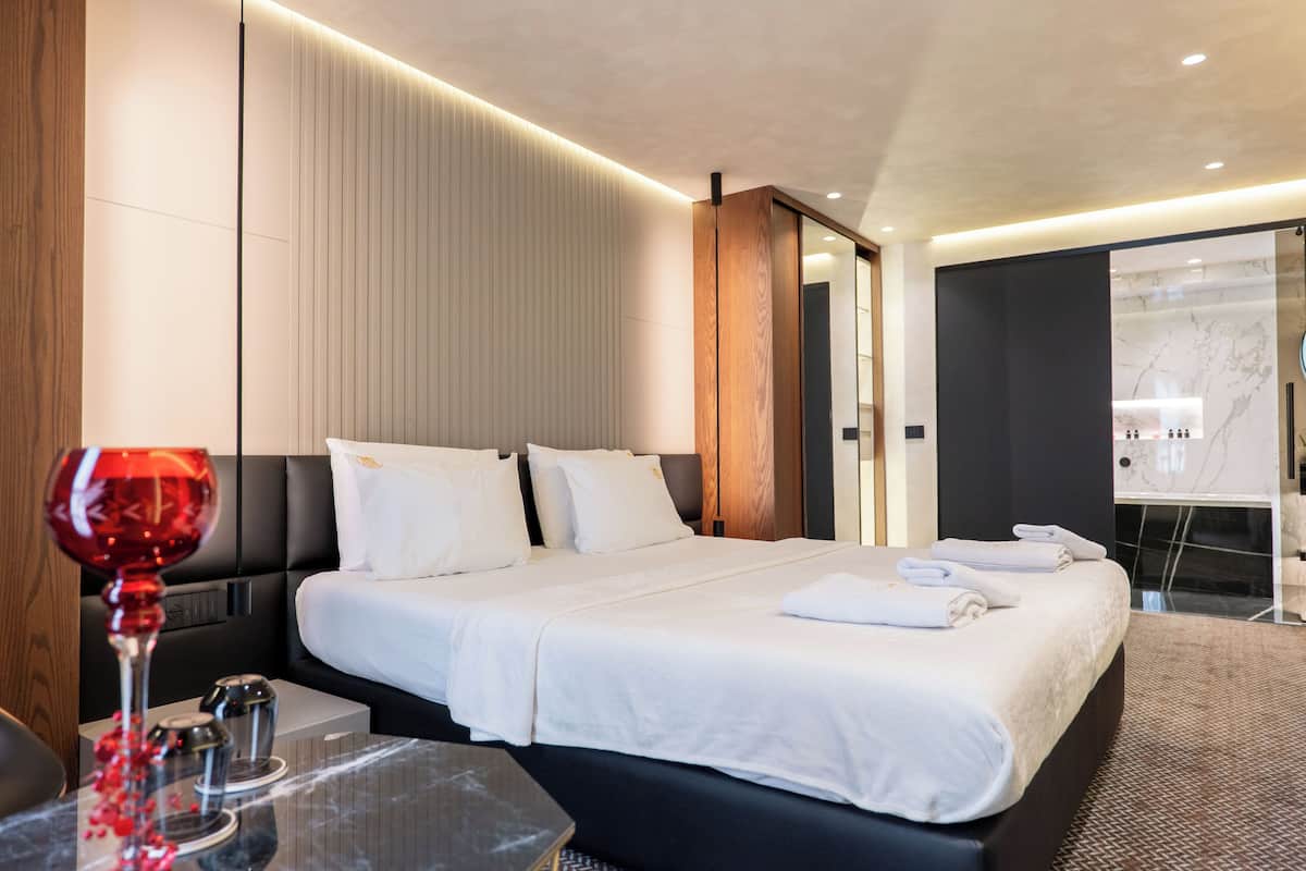 A modern hotel room with a large bed, bedside tables, a glass of red wine, and a bathroom with a marble shower.