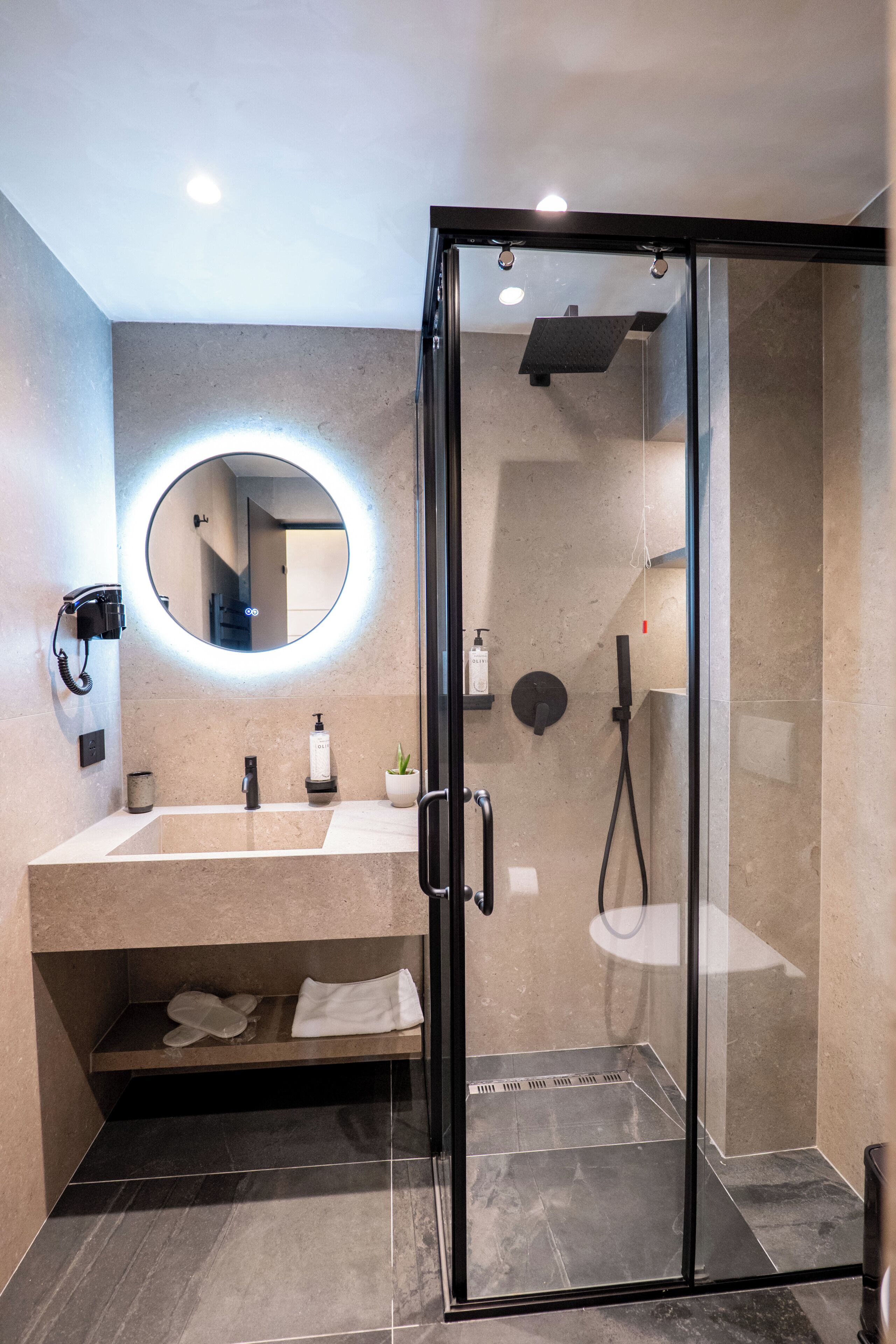 Une salle de bain moderne avec une douche à parois de verre, un grand miroir rond avec éclairage intégré, un lavabo avec un porte-serviettes et une petite plante en pot.