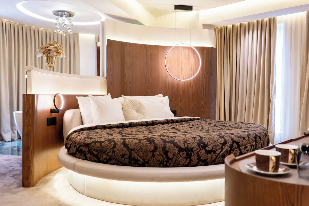 A modern hotel room with a circular bed, a bedside table with a cup and saucer, a round light fixture, and a wooden headboard.