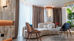 A modern living room with a curved sofa, a wooden chair, a glass coffee table, and a large plant. - Mattis Hotel (Tirana)