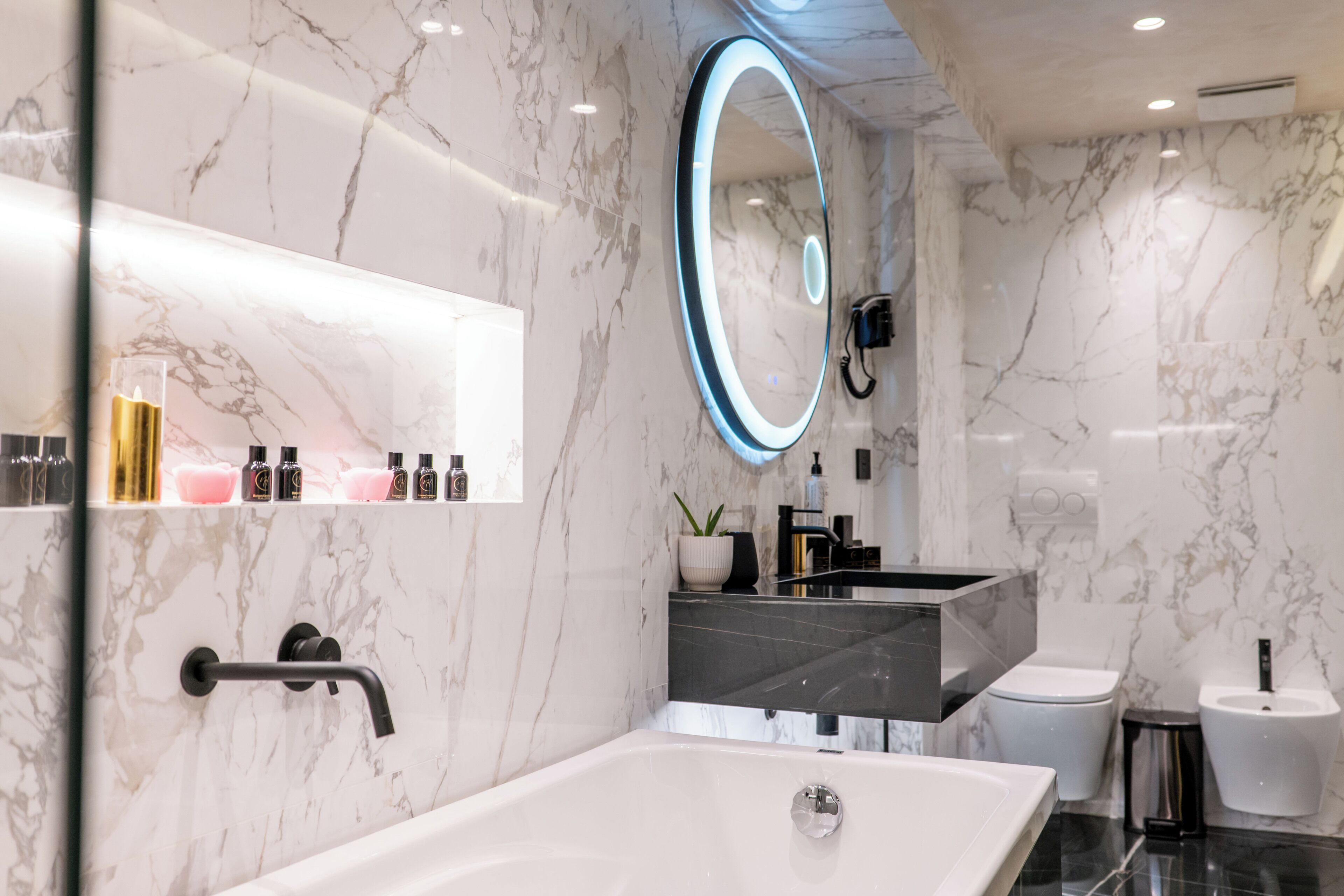 A modern bathroom with a marble wall, a freestanding bathtub, a vanity with a mirror, and a toilet.