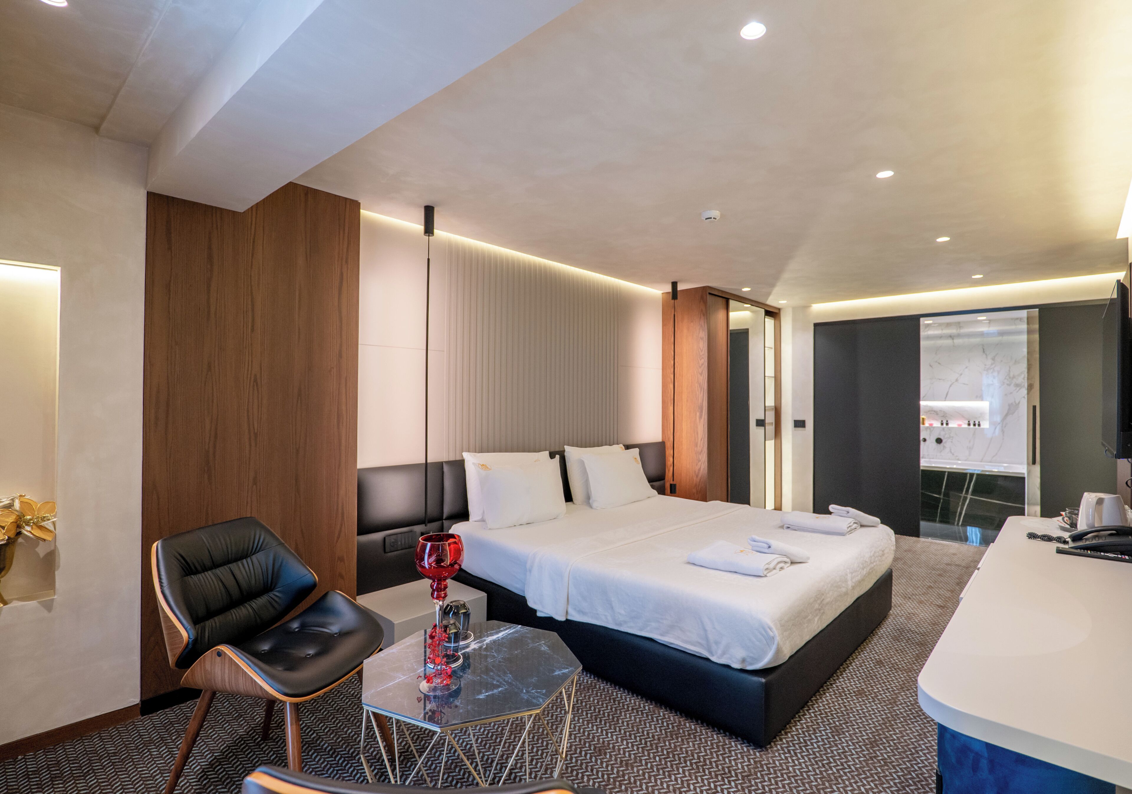 A modern hotel room with a large bed, a desk, a chair, and a small table with a red object.