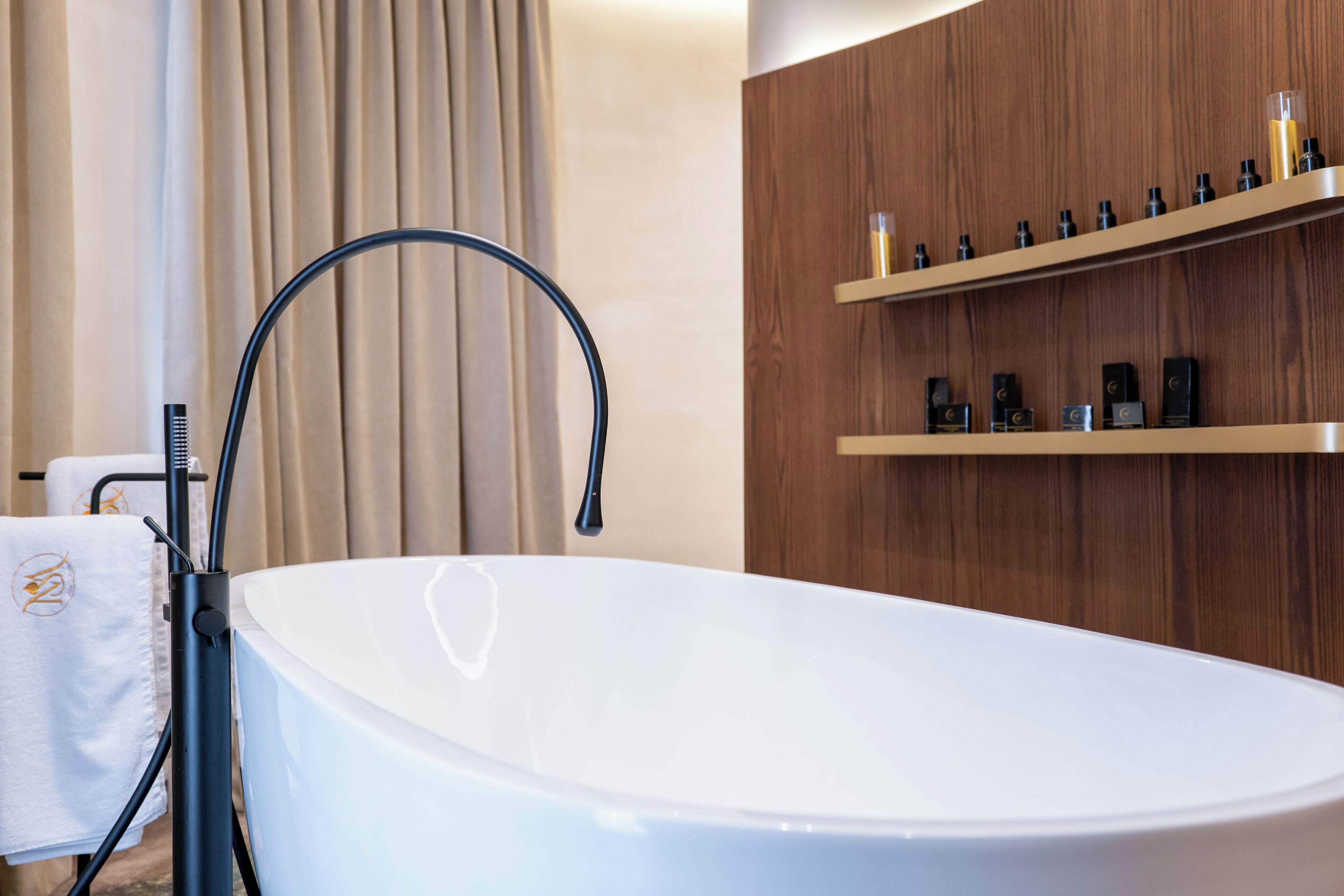 A modern bathroom with a freestanding bathtub, wooden shelving with toiletries, and a towel with a logo.