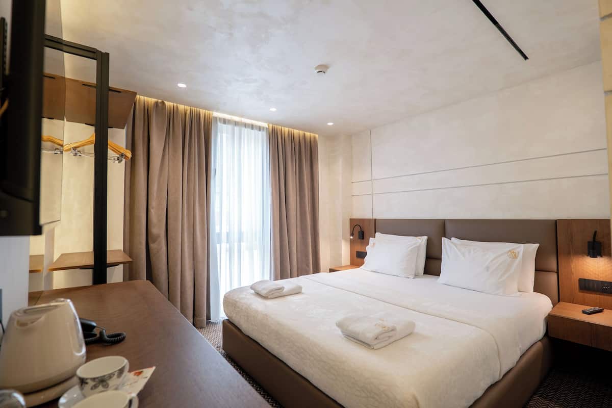 A hotel room with a large bed, a bedside table with a lamp, a desk with a telephone and a cup, and a mirror.