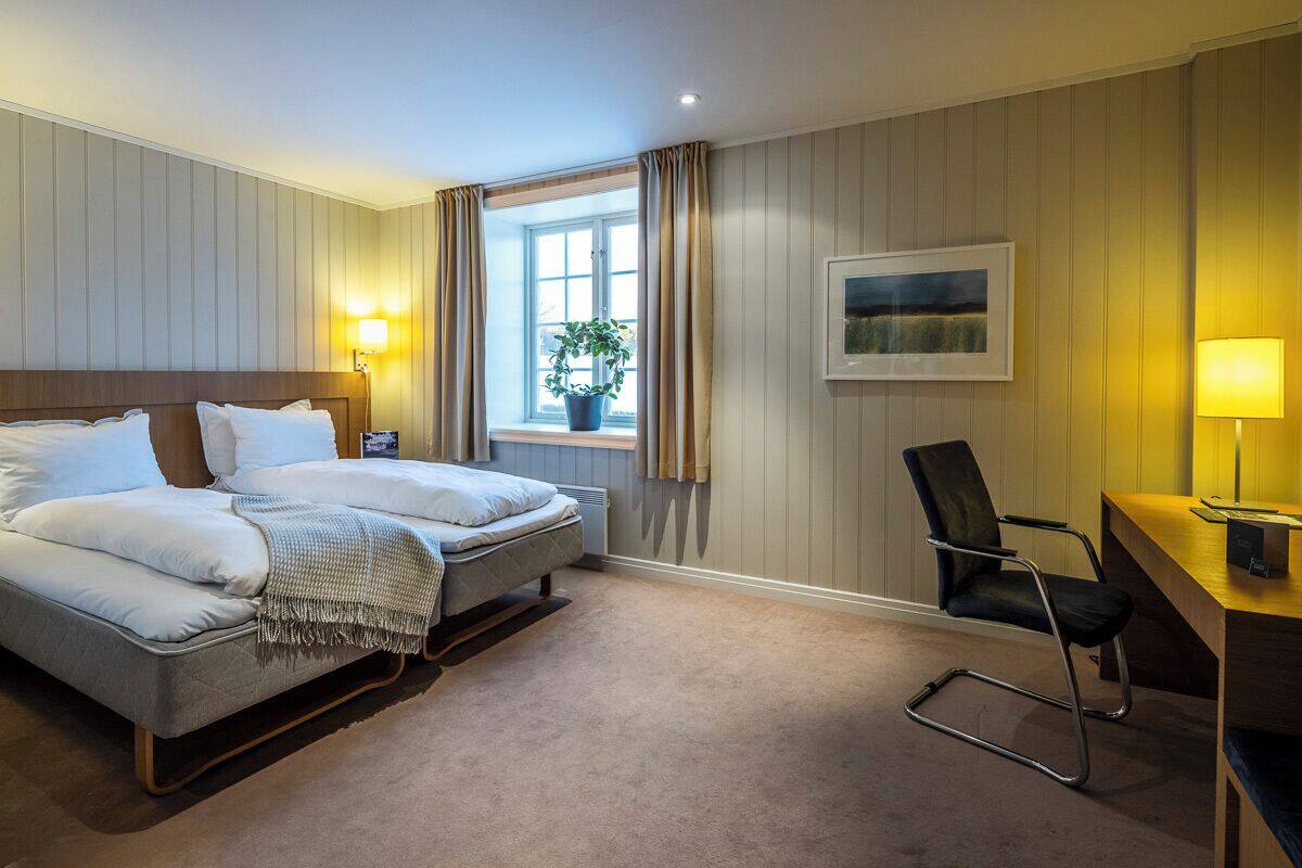 A hotel room with a bed, a desk, a chair, a window with curtains, a plant, and a painting on the wall.