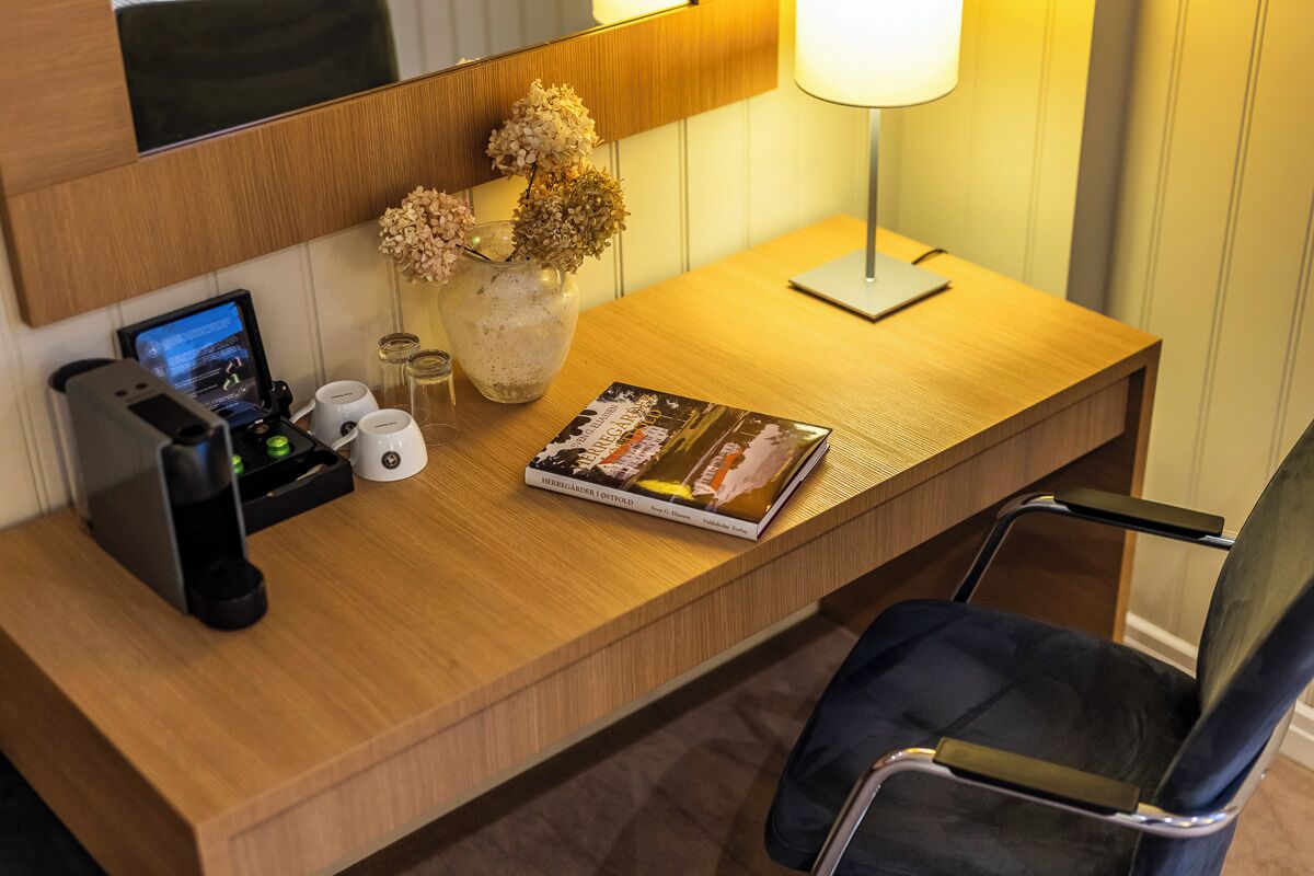 Un escritorio de hotel con una computadora portátil, un libro, un florero con flores secas, una lámpara y una cafetera.