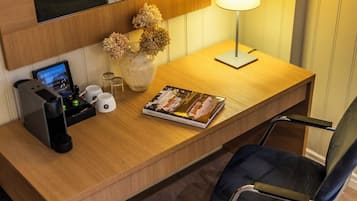 Un escritorio de hotel con una computadora portátil, un libro, un florero con flores secas, una lámpara y una cafetera.