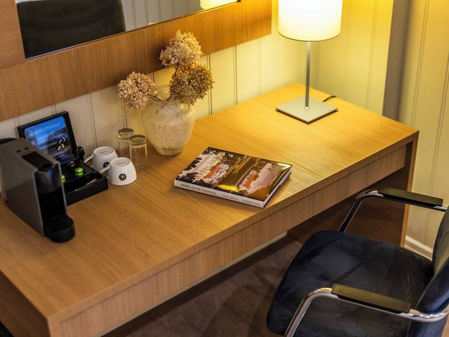 Un escritorio de hotel con una computadora portátil, un libro, un florero con flores secas, una lámpara y una cafetera.