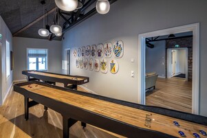 Una mesa de billar con superficie de madera y patas negras, una pared con varios escudos heráldicos, y una habitación con piso de madera y una puerta visible.