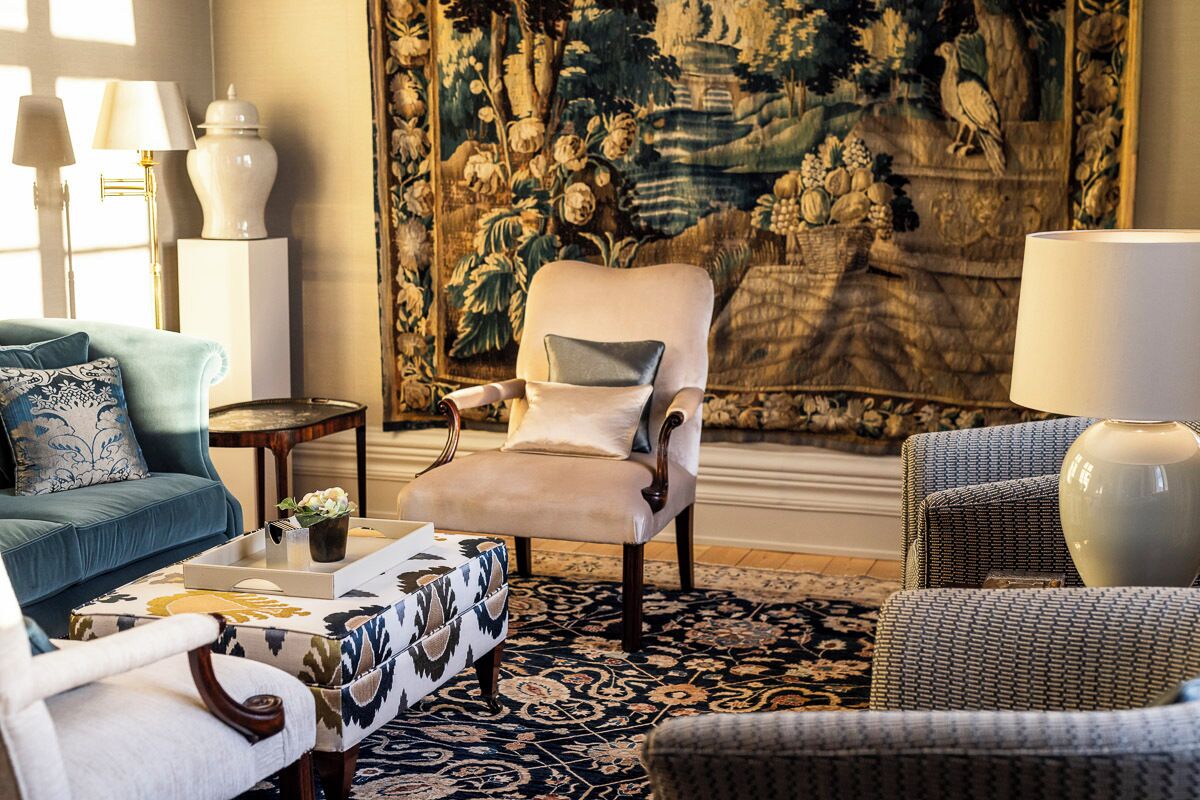 A living room with a blue sofa, a patterned ottoman, a beige armchair, and a large tapestry on the wall.