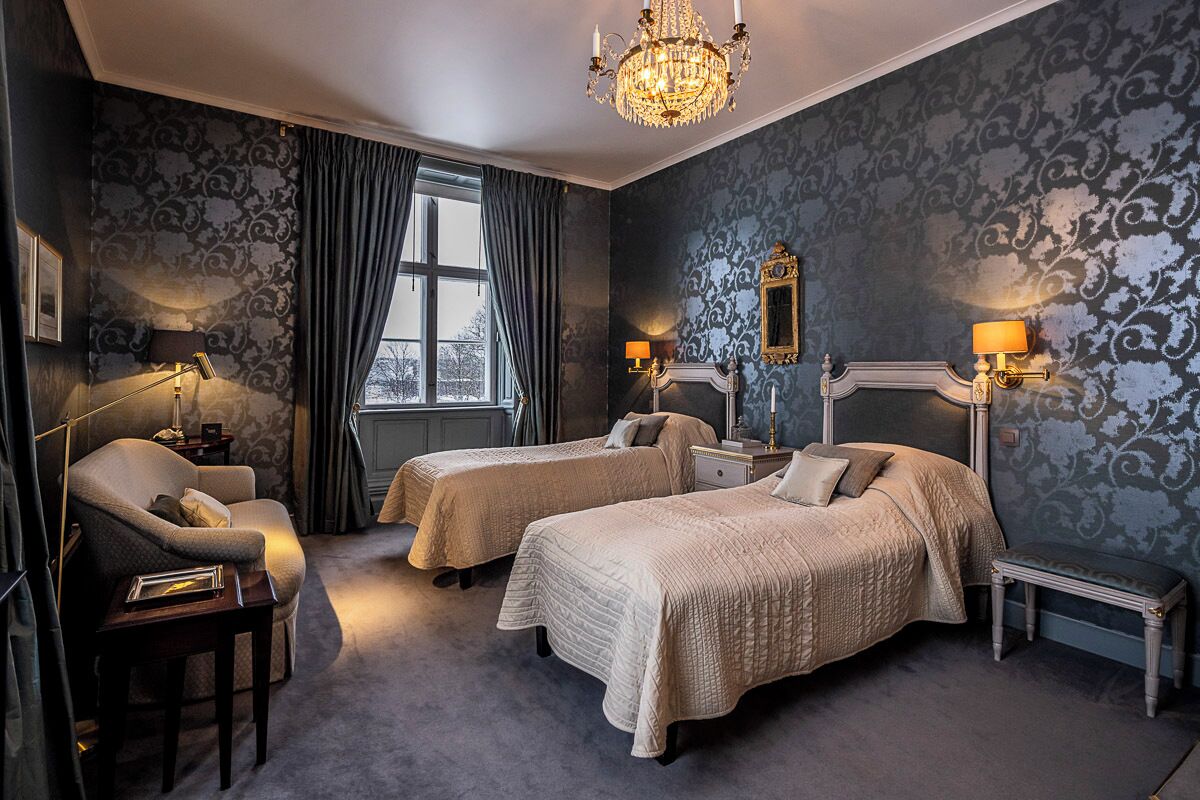 A hotel room with two beds, a chandelier, a window with curtains, and a mirror on the wall.