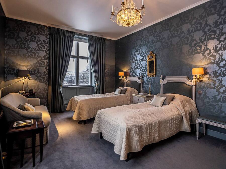 A hotel room with two beds, a chandelier, a window with curtains, and a mirror on the wall.