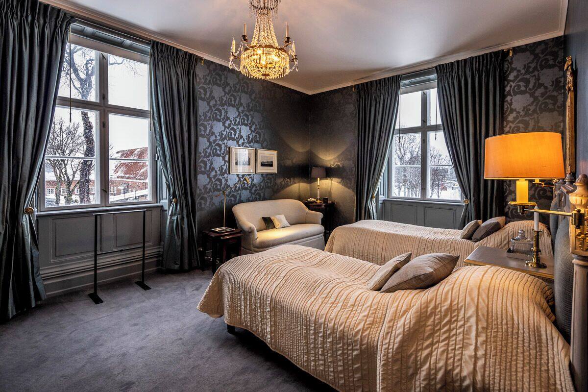 A hotel room with two beds, a chandelier, and large windows with curtains.